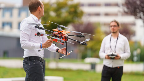 Photo of two IDiAL employees standing opposite each other on a lawn. One of them is holding a drone, the other is holding the controller for controlling the drone.