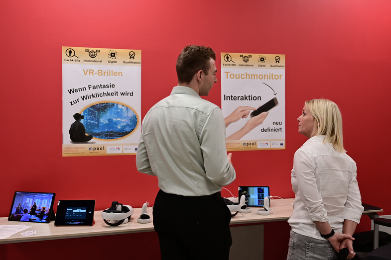 Station aus zwei Tischen vor einer roten Wand mit Postern. Zwei Personen unterhalten sich. Auf den Tischen stehen Tabletts und VR-Brillen. Text auf den Postern: „VR-Brillen - Wenn Fantasie zur Wirklichkeit wird“ und „Touchmonitor - Interaktion neu definiert“.