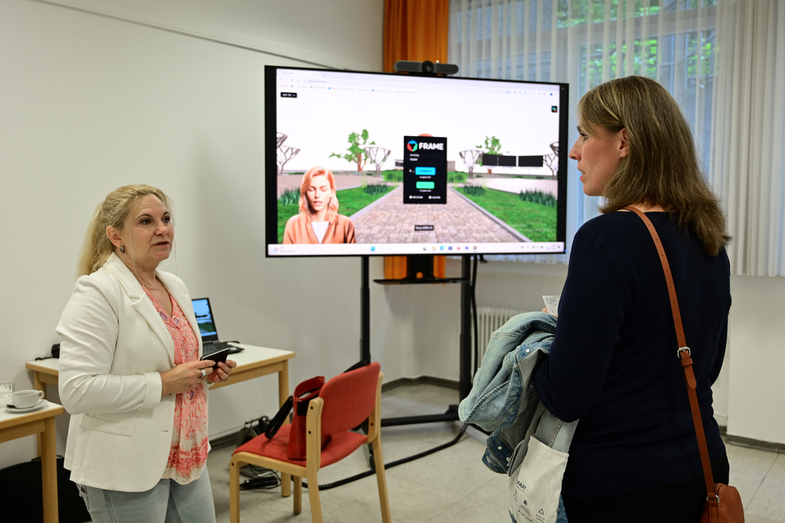 Zwei Personen unterhalten sich vor einem großen Fernseher mit Kamera. Auf dem Fernseher ist die Anmeldeseite zur Metaversum-Plattform mit eingeblendeter KI-Assistentin zu sehen.