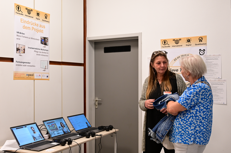 Informationsinsel mit drei Notebooks, die verschiedene Formulare mit eingeblendeten Assistenzsystemen zeigen. Es sind Headsets angeschlossen. Ein Poster mit dem Text „Eindrücke aus dem Projekt - VR-Brillen, wenn Fantasie zur Wirklichkeit wird - Innerbetriebliche Workshops: Pflege neu denken & direkt mitmachen - Formulargenerator, erstellen statt verzweifeln“. Zwei Personen stehen vor den Tischen und unterhalten sich.
