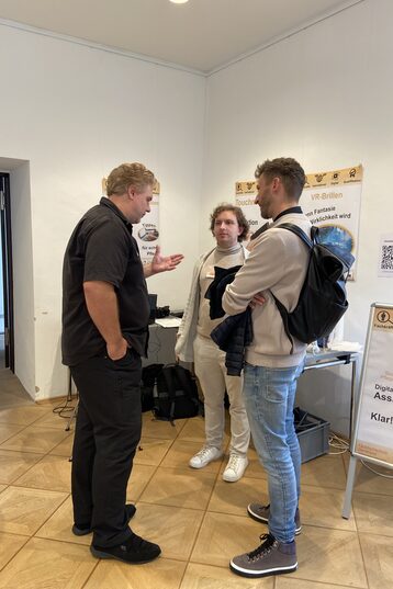 Standing at a table, Ansgar Rahmacher, Marcel Mitas and Leon Sachweh present assistance systems for the health and care sector.