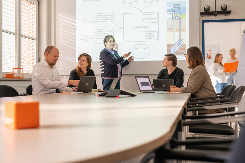 Photo of a meeting situation with 5 people. 4 are sitting at laptops and one woman is presenting standing up. 2 women are recognizable in the background in another room.