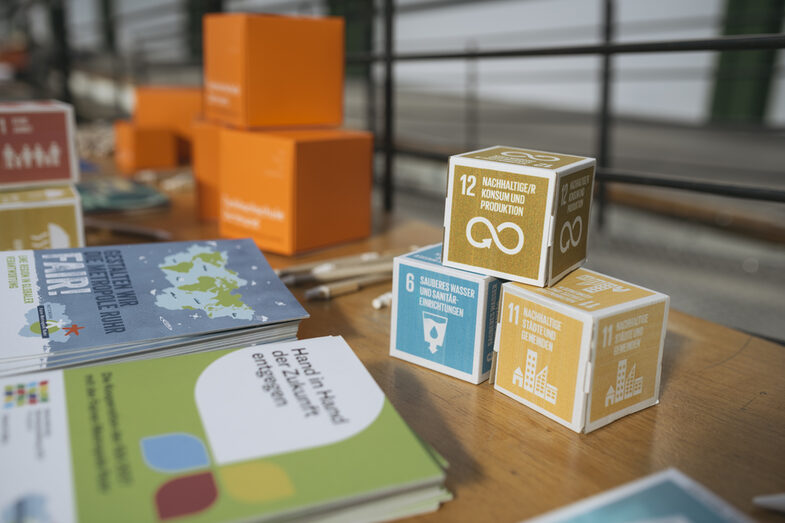 Auf einem Tisch liegen die SDG Würfel mit den Nachhaltigkeitszielen 6, 11, 12. Weiter hinten im Bild kann man die orangenen FH Würfel erkennen.