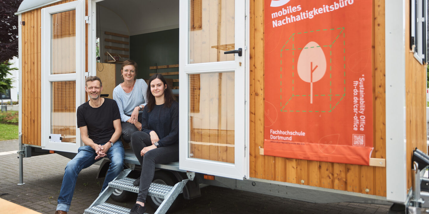 Man sieht 3 Personen, die in einem Bauwagen sitzen, welchen man über eine kleine Treppe betreten. An dem Bauwagen ist ein großes Plakat befestigt auf dem „Soffi mobiles Nachhaltigkeitsbüro“ steht.