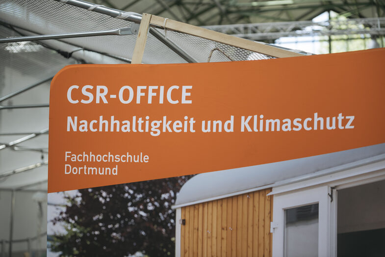 Auf dem Bild ist ein Ausschnitt von einer Werbewand des CSR-Office zu erkennen auf dem steht "CSR-Office Nachhaltigkeit und Klimaschutz, Fachhochschule Dortmund. Die Werbewand schein in einer Halle zu stehen, da man Dachträger im Hintergrund erkennen kann.