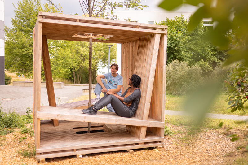 Photo of 2 students sitting and talking in a wooden cube equipped with seating. A tree is planted in the middle of the cube.