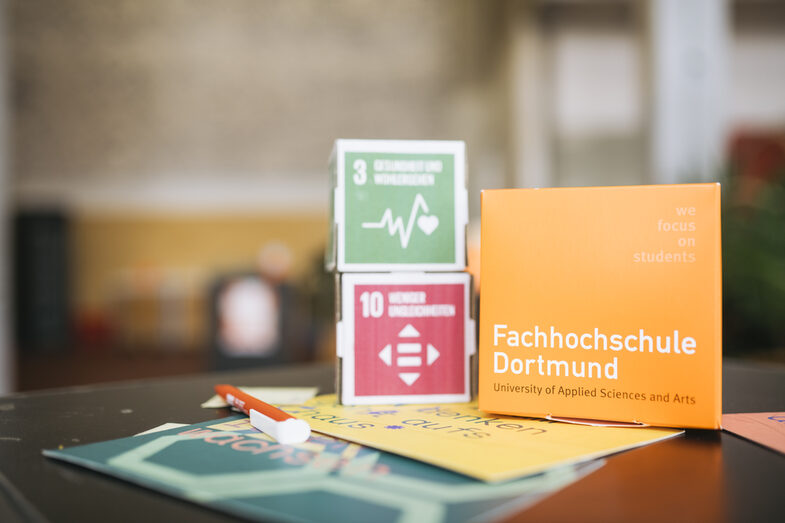 Auf dem Bild ist in Nahaufnahme ein FH Pappwürfel und zwei Würfel mit den Nachhhaltigkeitszielen 3 und 10 auf einem Tisch zu sehen. Vor den Pappwürfeln liegen Broschüren und Flyer