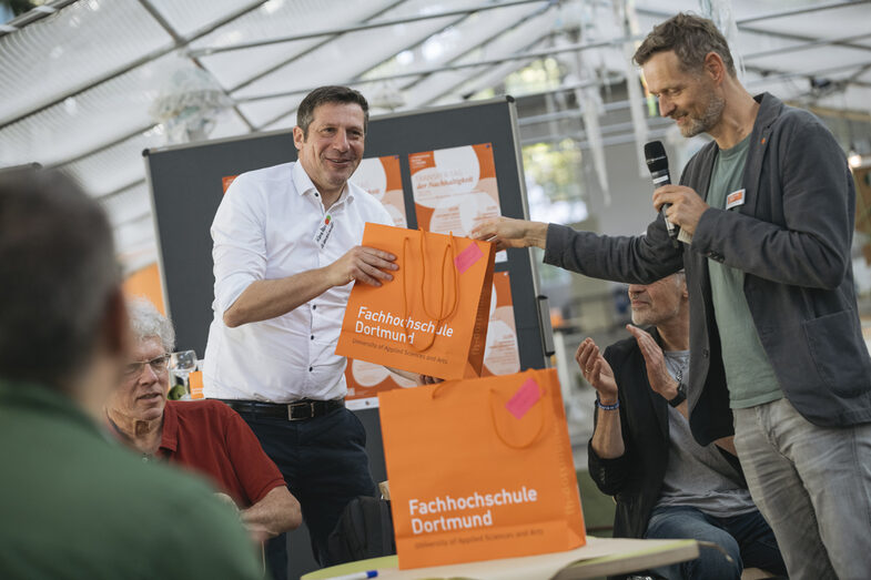 Auf dem Bild sind zwei Personen zu sehen. Die eine Person reicht der anderen Person eine orangene Tüte mit der Aufschrift Fachhochschule Dortmund. Die gebende Person hat ein Mikrofon in der Hand und spricht zu einem Publikum. Unscharfe Umrisse von Personen die dabei sitzen sind vorne links zu erkennen.