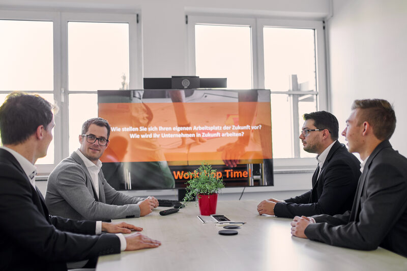 Four people are sitting at a table in a conference room. At the end of the table is a large television with a conference camera attached to it. A presentation is being played on the TV. The four people are discussing something.