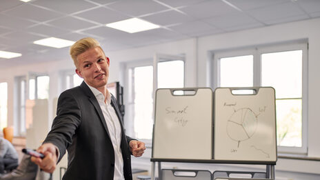 Hier steht eine Person vor einer Halterung mit drei kleineren Whiteboards und hat den Arm ausgestreckt. In seiner Hand hat er einen Board Marker, welchen er scheinbar einer anderen Person übergeben will, damit die jeweilige Person auch etwas auf die Whiteboards schreiben kann.