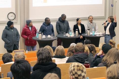 Sieben Personen an Stehtischen vor gut besetzten Hörsaal-Sitzreihen schauen einander an. Die Person ganz rechts spricht in ein Mikro und gestikuliert.