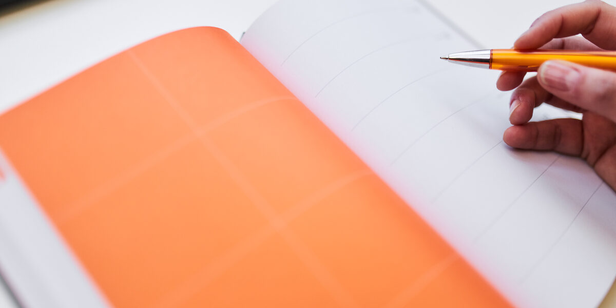 Photo of an open diary, the left-hand page is orange. Someone starts to write something in it with a ballpoint pen.