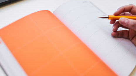 Photo of an open diary, the left-hand page is orange. Someone starts to write something in it with a ballpoint pen.