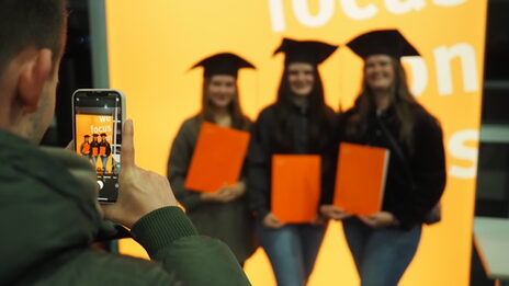 Graduates stand in front of a light wall and are photographed by a person with a cell phone