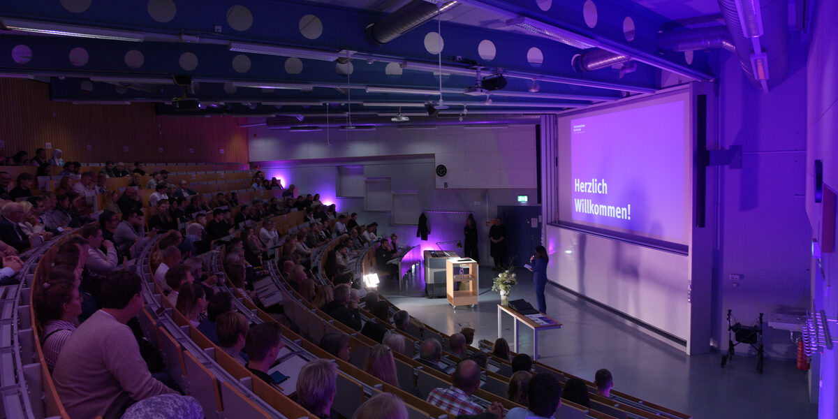 A packed lecture hall in which a woman is speaking at the front. The projection shows "Welcome!" in purple lighting.