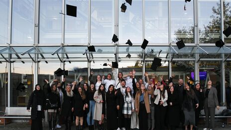 The graduates stand in front of the faculty building and throw their graduation hats in the air.