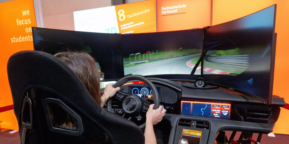 Eine Person sitzt im Fahrsimulator, steuert das Lenkrad und schaut auf drei Bildschirme.