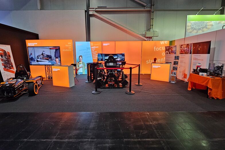 An exhibition stand is set up in a hall, with a racing car, a racing simulator and a table with technical elements in front of an orange illuminated wall.