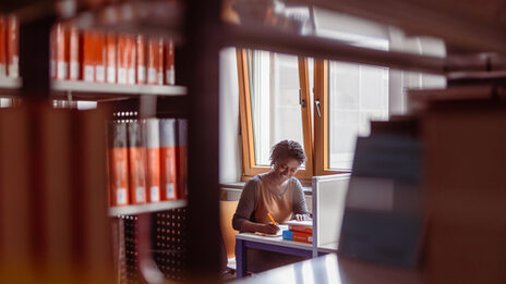 Foto von einer Studentin, die etwas schreibt. Der Blick der Kamera geht durch eine Lücke im Bücherregal hindurch zu dem Lernplatz der Studentin.