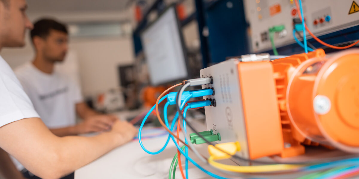 Foto von einem Elektromotor, im Hintergrund arbeiten 2 Studierenden am Computer des Prüfstandes.