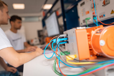 Photo of an electric motor, in the background 2 students working on the test bench computer.
