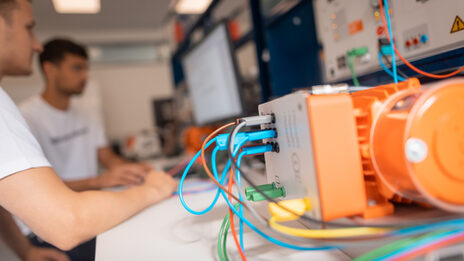Photo of an electric motor, in the background 2 students working on the test bench computer.