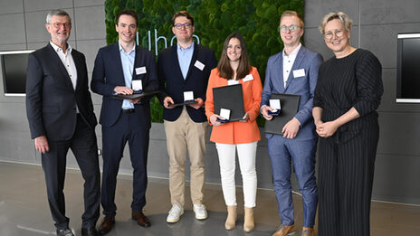 Sechs Personen stehen in einer Reihe nebeneinander, die mittleren vier halten eine Mappe und ein Schachtel mit einer Medaille vor sich.