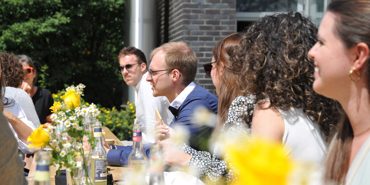 View of the long table decorated with flowers set up in front of the building, where former students are chatting animatedly.