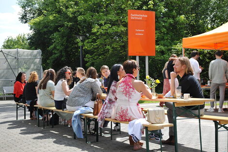 Blick auf den vor dem Gebäude aufgebauten Tisch, an dem sich die ehemaligen Studierenden angeregt unterhalten, mit orangefarbenen FH-Pavillion im Hintergrund.