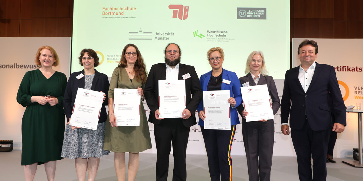 Sieben Personen stehen nebeneinander auf einer Bühne, fünf halten ein Zertifikat vor sich. Im Hintergrund auf einer Leinwand zu sehen sind die Logos der Fachhochschule Dortmund, der TU Berlin, der TU Dresden, der Uni Münster und der Westfälischen Hochschule.