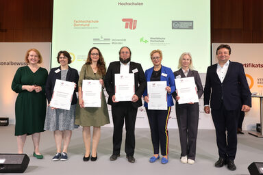 Sieben Personen stehen nebeneinander auf einer Bühne, fünf halten ein Zertifikat vor sich. Im Hintergrund auf einer Leinwand zu sehen sind die Logos der Fachhochschule Dortmund, der TU Berlin, der TU Dresden, der Uni Münster und der Westfälischen Hochschule.