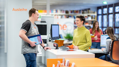 Photo of a member of staff behind the library counter, she is in conversation with the student standing opposite her, who is holding books. In the background, another student is talking to a member of staff.