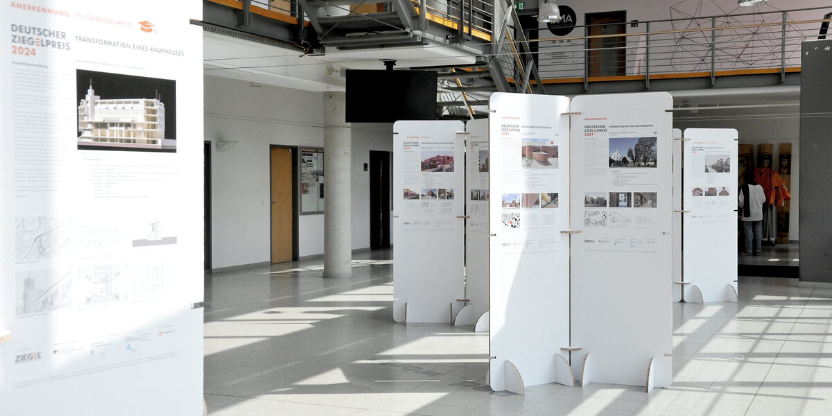 Ausstellung im Foyer des Fachbereichs mit freistehenden weißen Tafeln, auf denen Architekturprojekte zum Deutschen Ziegelpreis 2024 präsentiert werden.
