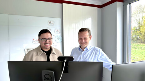 Two people stand behind two monitors and look at them.