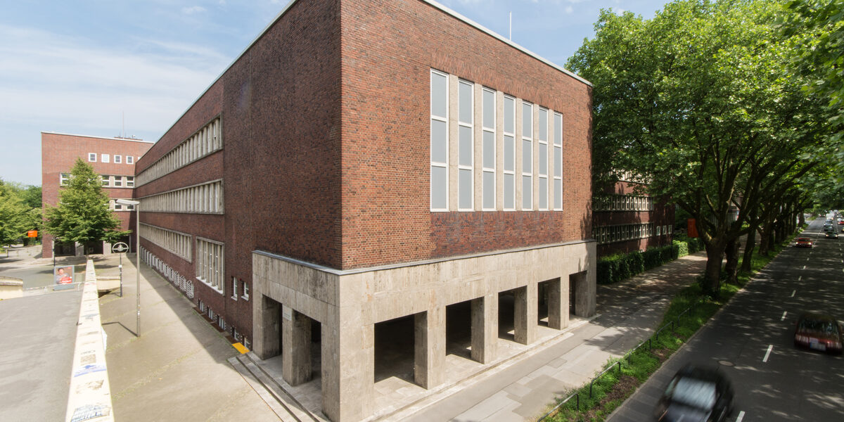 Foto mit Blick auf Teile eines Gebäudes der Fachhochschule Dortmund am Max-Ophüls-Platz mit Vorplatz und Gehweg sowie Straße mit Autos daneben.