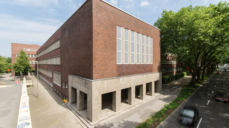 Photo with a view of parts of a building of the Fachhochschule Dortmund at Max-Ophüls-Platz with forecourt and sidewalk as well as street with cars next to it.