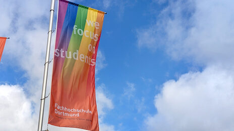 A Fachhochschule Dortmund flag with a rainbow flies against a blue sky.