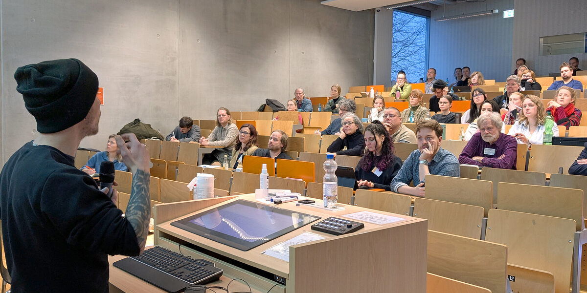 Eine männlich gelesene Person spricht zu Menschen in einem Hörsaal.