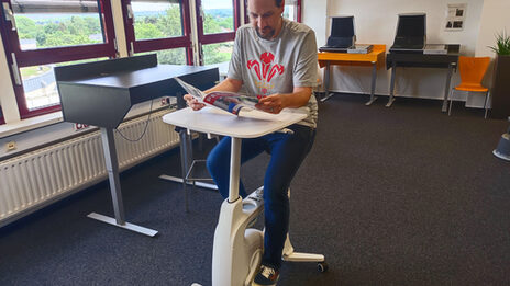 In the middle of a room is a bicycle-like device, albeit without wheels and handlebars, but with a small table. A person sits on the device, pedals and reads a magazine.