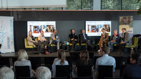 8 people are sitting on a stage during a panel discussion. The audience is sitting in front of them. Prof. Dr. Sabine Sachweh is sitting on the far left of the stage.