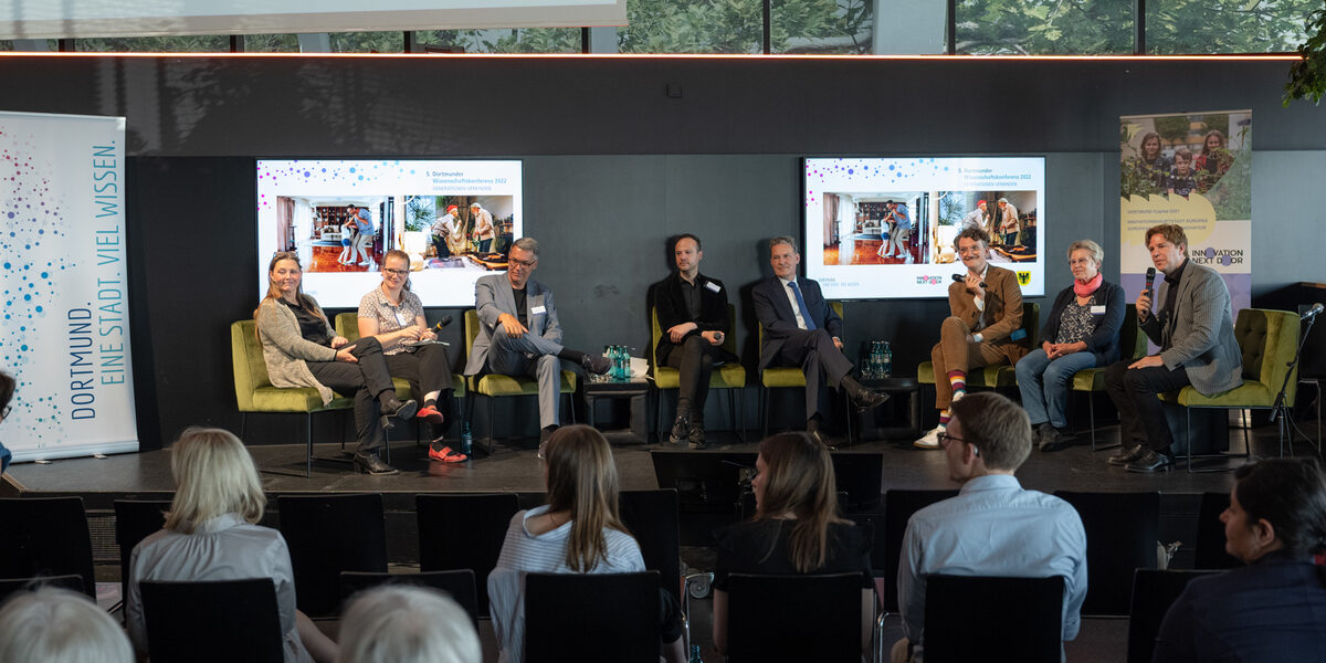 8 people are sitting on a stage during a panel discussion. The audience is sitting in front of them. Prof. Dr. Sabine Sachweh is sitting on the far left of the stage.