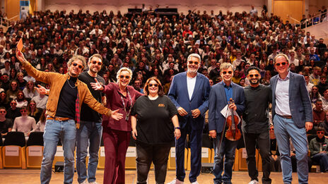 Several people with sunglasses are standing on a stage. A full hall can be seen in the background.