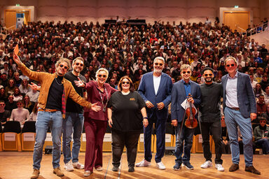 Several people with sunglasses are standing on a stage. A full hall can be seen in the background.