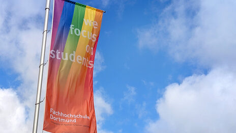 A Fachhochschule Dortmund flag with a rainbow flies against a blue sky