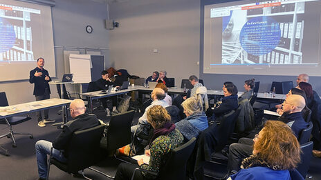 In a large, functional room, a person stands on the left at a high table with a laptop and gesticulates, while 18 people sit at tables spread around the room facing him. Projections can be seen on the wall to the right and left.