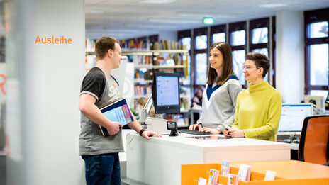 Photo of two staff members standing behind the library counter and talking to the student opposite them, who is holding books.