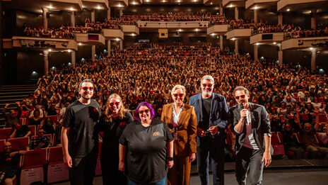 Several people wearing sunglasses on stage in the opera house. In the background, the first-semester students in the stands make "la Ola".