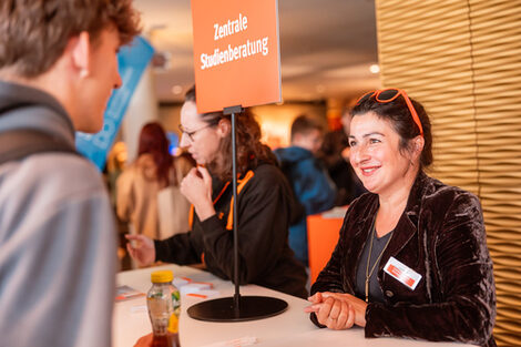 Zwei Personen sprechen miteinander. Auf dem Tisch steht ein Schild mit der Aufschrift "Zentrale Studienberatung".