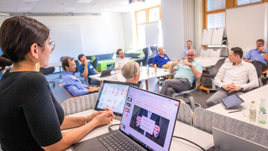 Foto von einer Veranstaltung in einem Seminarraum. Im Fokus ist eine junge Dame am Rednerpult, wo auch ein Laptop draufsteht, diese lauscht gespannt dem Redner*in in der Gruppe.