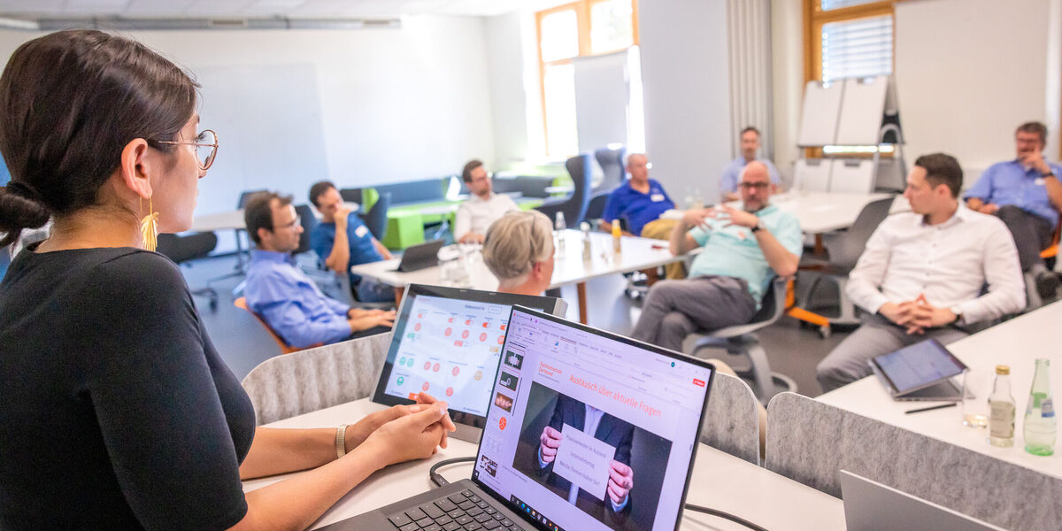 Foto von einer Veranstaltung in einem Seminarraum. Im Fokus ist eine junge Dame am Rednerpult, wo auch ein Laptop draufsteht, diese lauscht gespannt dem Redner*in in der Gruppe.